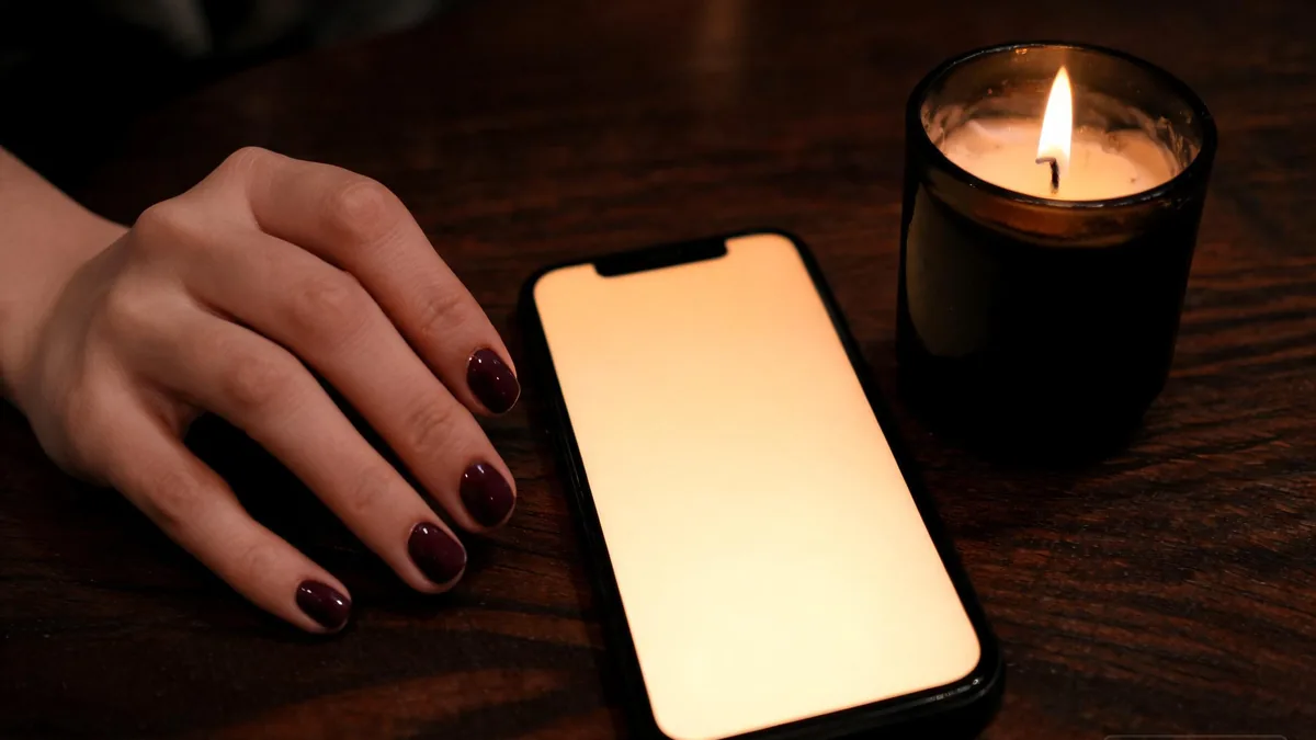 Goth AI girlfriend companion through a chat app, a soft warm glow on a dark wood table with a black candle and deep plum nails resting nearby