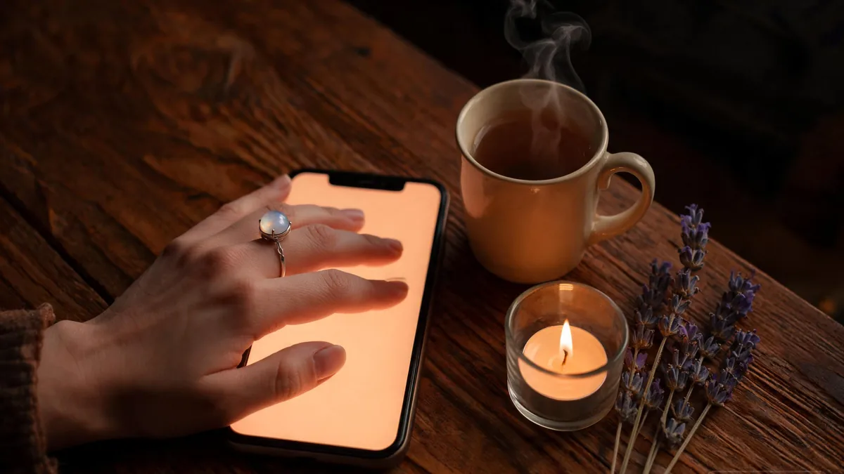 Witch AI girlfriend companion experienced through a phone, a hand with a delicate moonstone ring resting beside a mug of herbal tea, a candle, and sprigs of dried lavender